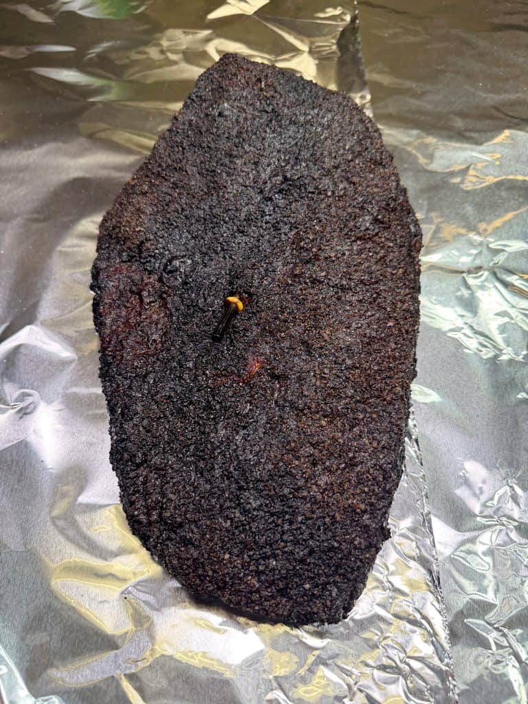 A whole smoked brisket with a dark, crusty bark rests on a sheet of aluminum foil. A meat probe is inserted into the center of the brisket.
