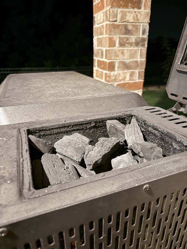 A close-up of a metal grill or smoker with its hatch open, showing several pieces of charcoal inside. A brick column and a dark outdoor background are visible.