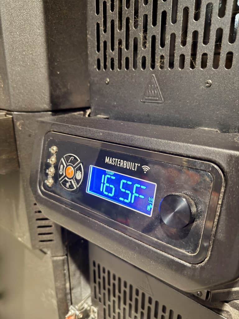 Close-up of a Masterbuilt digital smoker control panel displaying a temperature of 165.5°F in bright blue numbers, with various control buttons and a dial on a black metal surface.