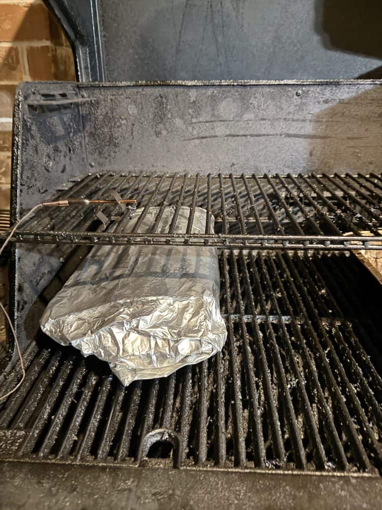 A foil-wrapped item is placed on the lower rack inside a black, open barbecue smoker. The interior of the smoker is slightly greasy with visible grill marks on the racks.