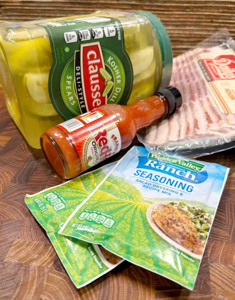 A jar of kosher dill pickle spears, a pack of bacon, two Hidden Valley Ranch seasoning packets, and a bottle of Franks RedHot sauce arranged on a wooden surface.