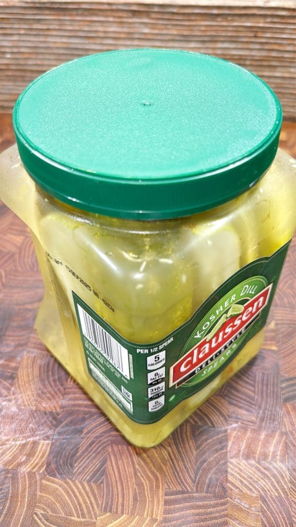 A large square plastic jar of Claussen Kosher Dill pickles with a green lid sits on a wooden surface. The pickles are visible through the clear container.