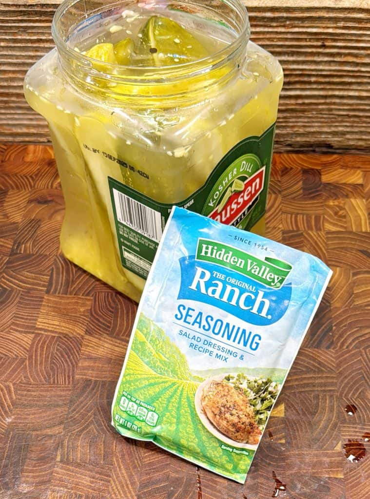 A packet of Hidden Valley Ranch seasoning lies in front of an open jar of Vlasic Kosher Dill pickles on a wooden surface.
