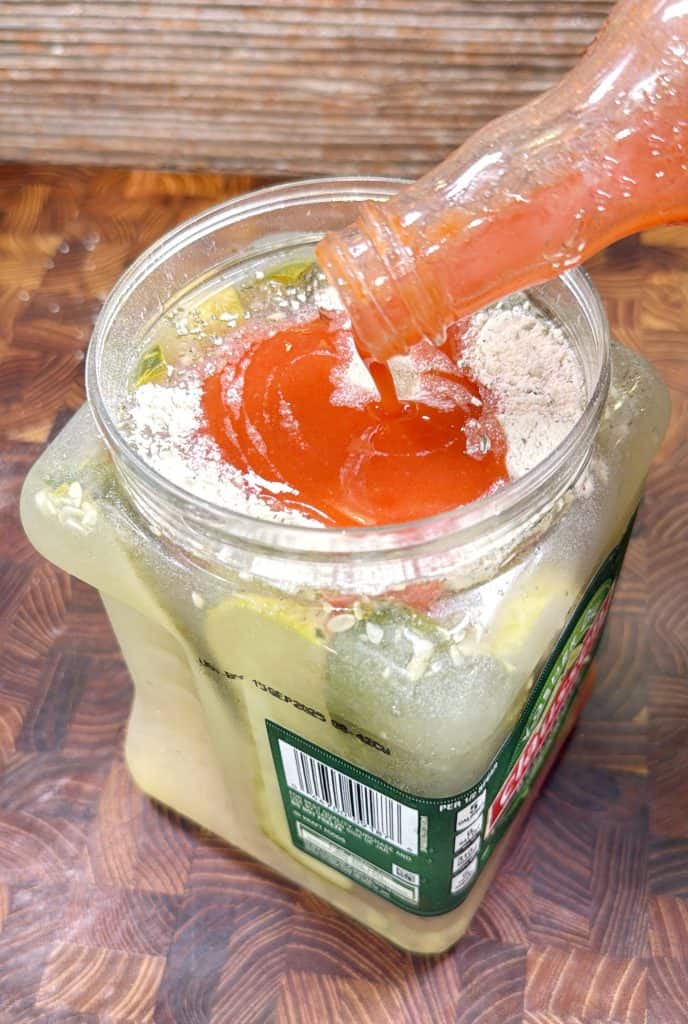 A bottle of hot sauce is being poured into a large plastic jar filled with pickles, juice, and seasonings, sitting on a wooden surface.