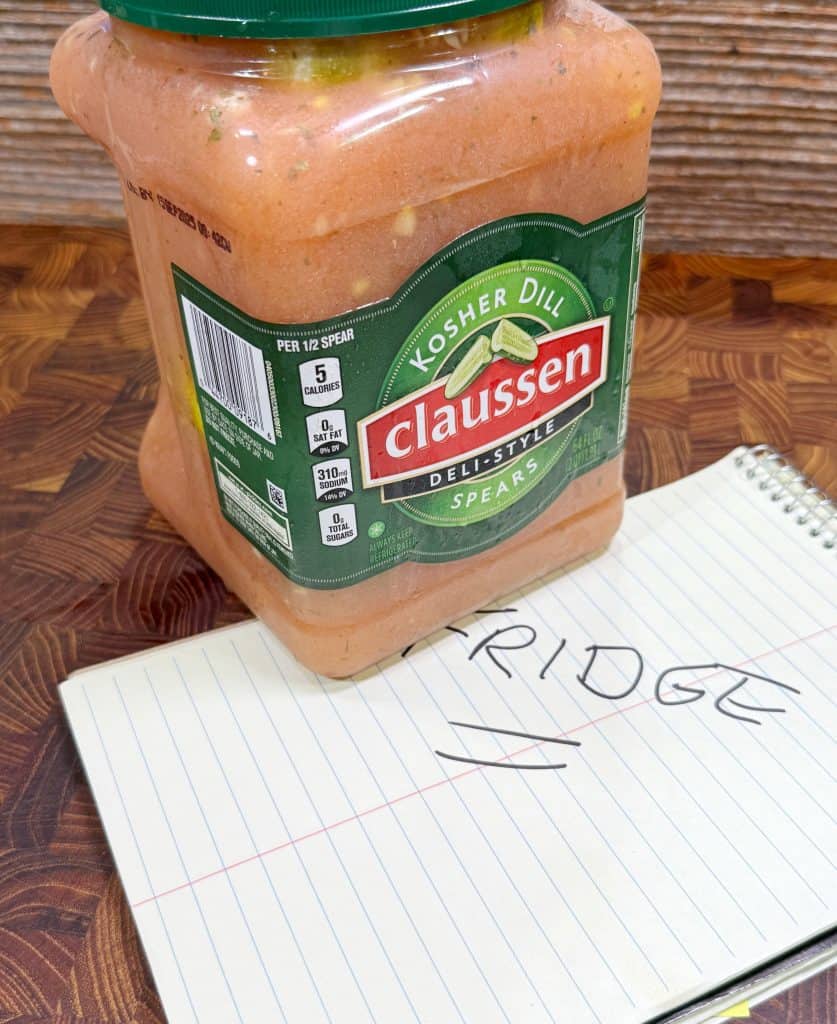 A jar of Claussen Kosher Dill pickle spears sits on a wooden surface next to a notepad with FRIDGE written in large letters.