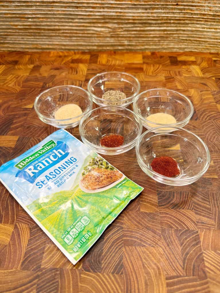 Six small glass bowls containing various spices are arranged on a wooden surface next to a packet of Hidden Valley Ranch Seasoning. The spices appear to be chili powder, paprika, onion, garlic, and herbs.