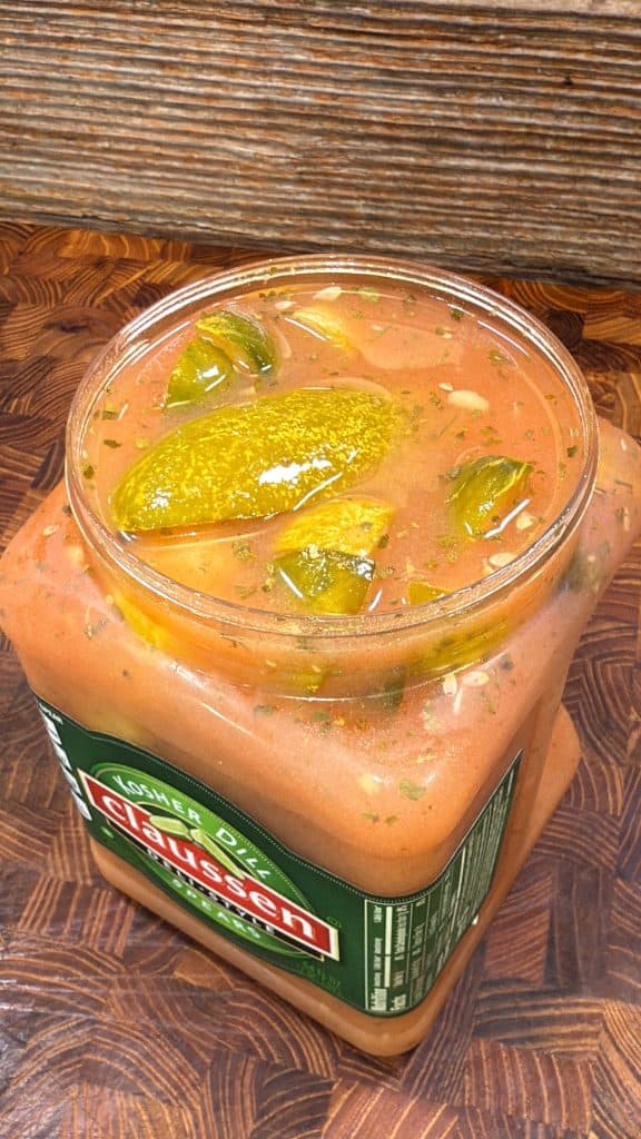 An open jar of Claussen pickles filled with reddish brine, herbs, and several pickle spears, sitting on a wooden surface. The jar is full, with liquid and pickles nearly overflowing the top.