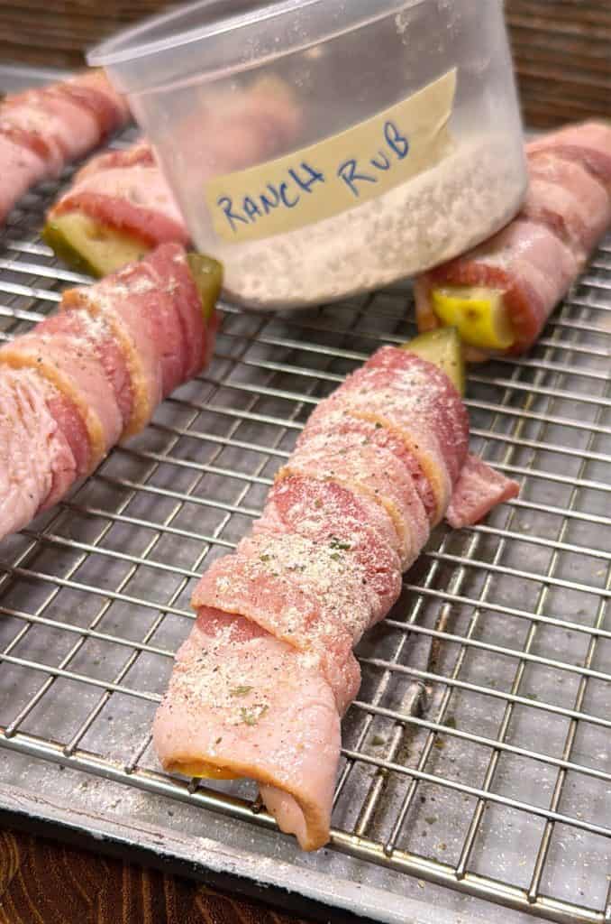 Close-up of bacon-wrapped pickles sprinkled with ranch seasoning on a wire rack, with a plastic container labeled RANCH RUB in the background.