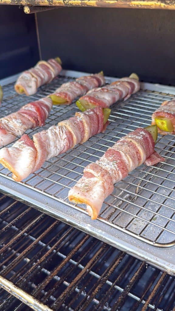 Bacon-wrapped skewers with green peppers are arranged on a wire rack inside a smoker or grill, being cooked with visible seasoning on top.