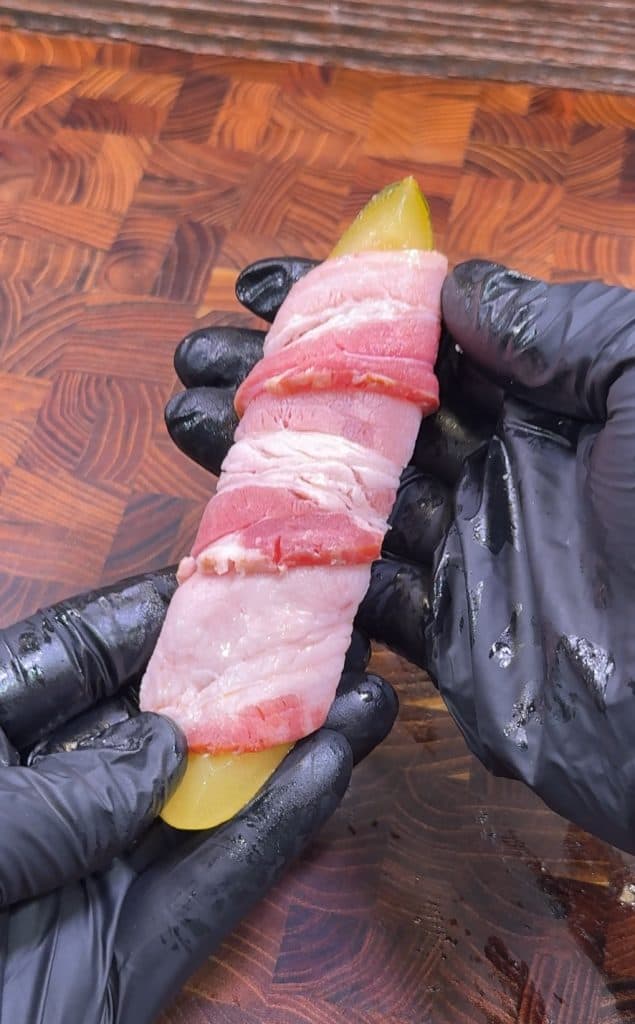 A person wearing black gloves holds a pickle spear wrapped tightly with raw bacon over a wooden cutting board.