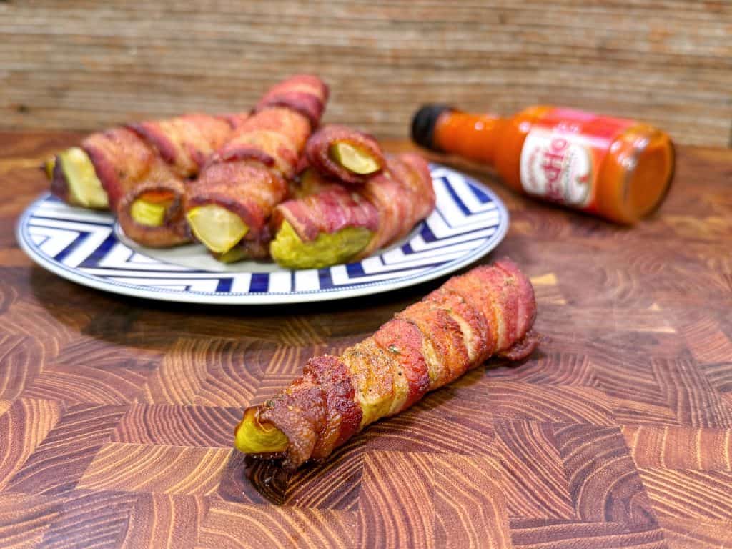 Bacon-wrapped pickles are displayed on a patterned plate, with one piece in the foreground on a wooden surface. A bottle of hot sauce is lying on its side in the background.
