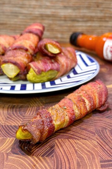 Bacon-wrapped pickles are displayed on a patterned plate, with one piece in the foreground on a wooden surface. A bottle of hot sauce is lying on its side in the background.