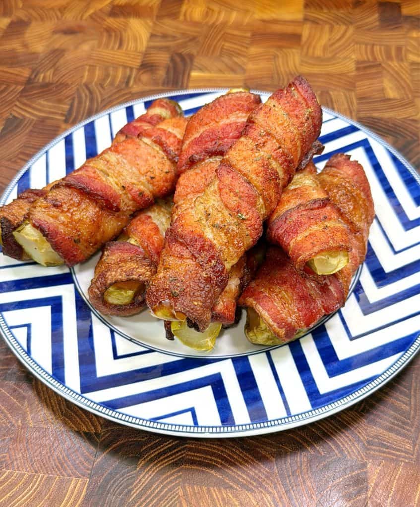 A plate with a blue and white zigzag pattern holds several bacon-wrapped jalapeño poppers arranged in a pile on a wooden surface. The bacon looks crispy and golden brown.