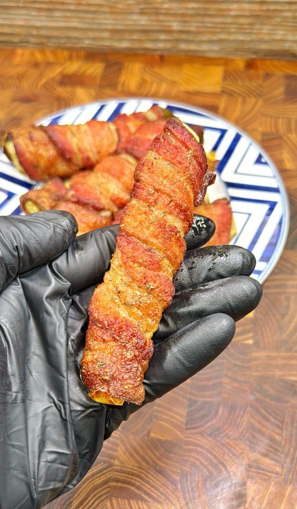 A gloved hand holds a bacon-wrapped food item, with more bacon-wrapped pieces on a patterned plate in the background, all set on a wooden surface.