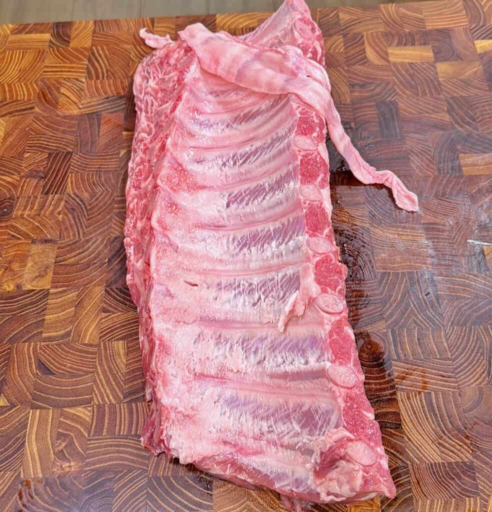 A slab of raw pork ribs with some attached meat and membrane sits on a wooden cutting board with a distinct checkered pattern.