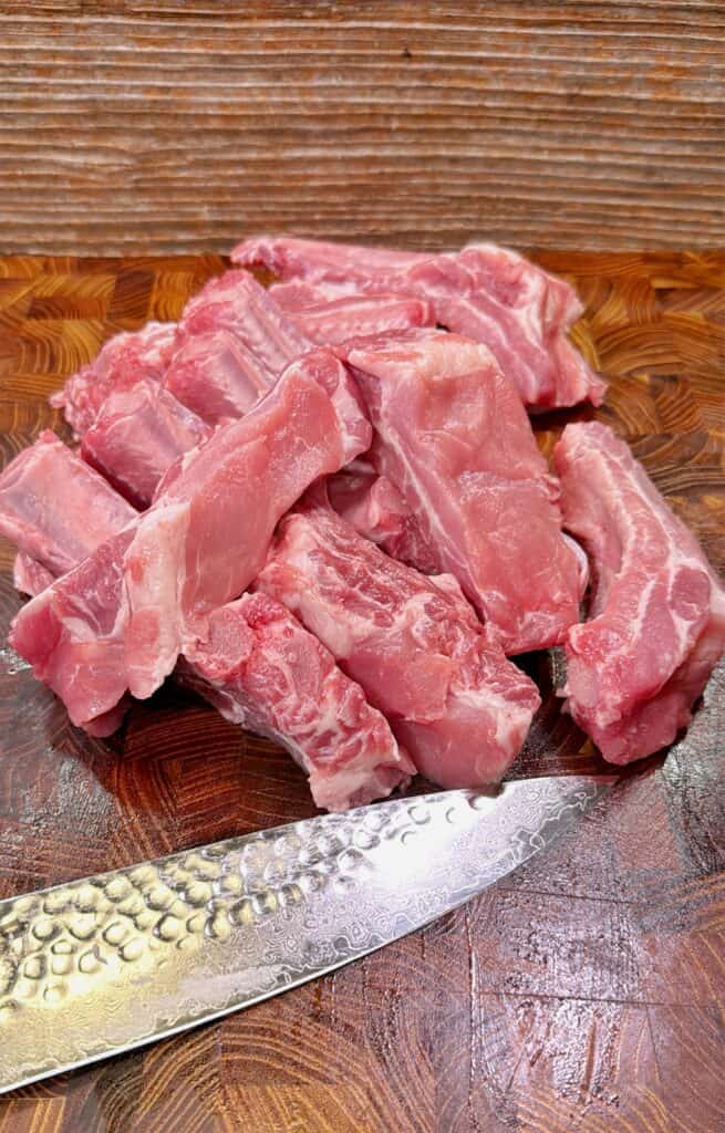 Raw pieces of pork ribs are stacked on a wooden cutting board with a silver, textured chef’s knife placed in front of them. The background shows wood paneling.