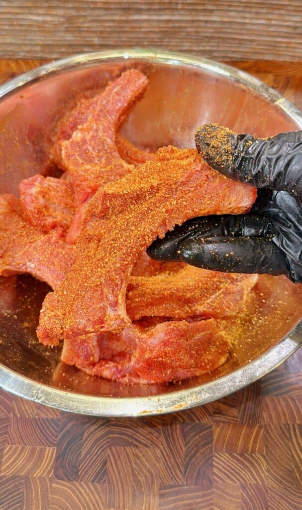 A gloved hand holds a raw pork rib coated in red seasoning over a metal bowl filled with more seasoned ribs, resting on a wooden surface.