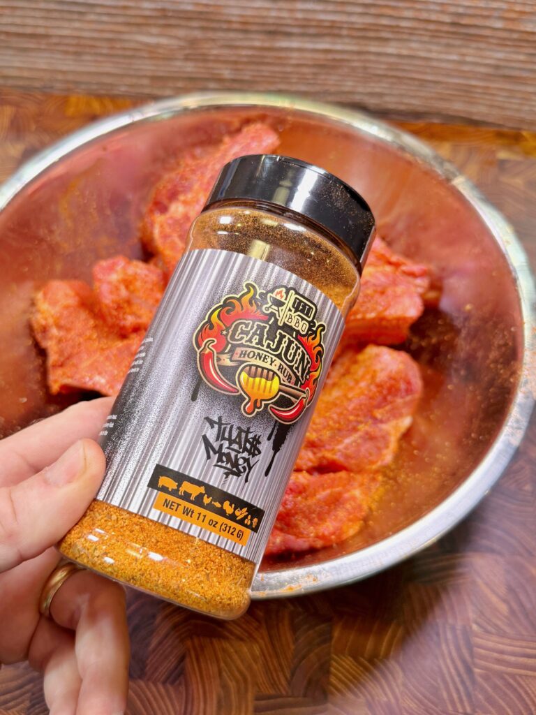 A hand holds a bottle of Cajun Honey Heat seasoning in front of a metal bowl filled with raw, seasoned meat on a wooden surface.