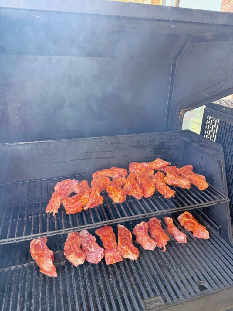Seasoned meat strips cooking on the grates of a barbecue grill with smoke rising, and the grill lid partially open.