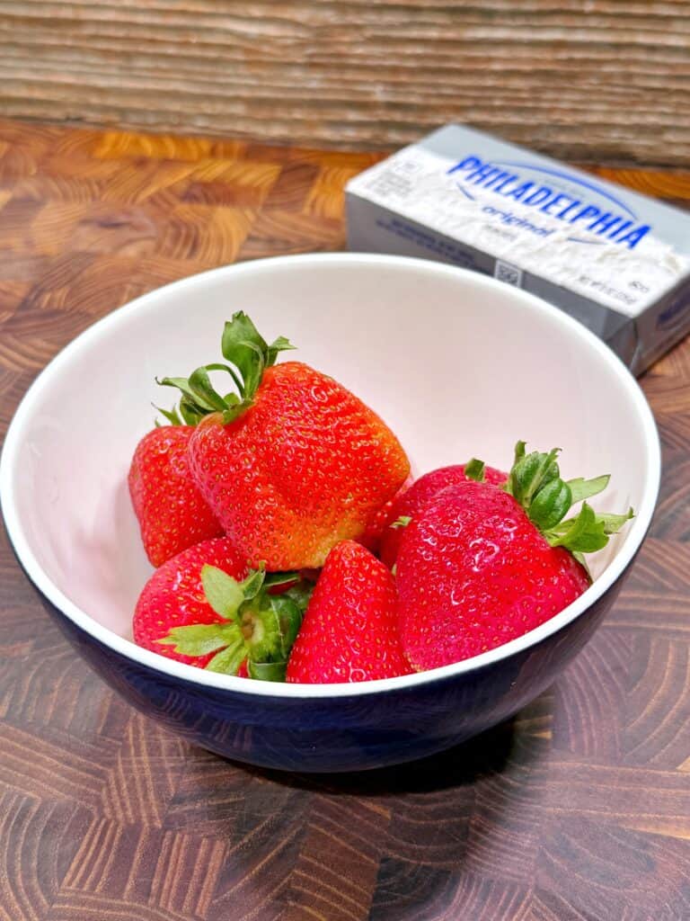 A bowl filled with fresh strawberries sits on a wooden surface, with a package of Philadelphia cream cheese in the background.