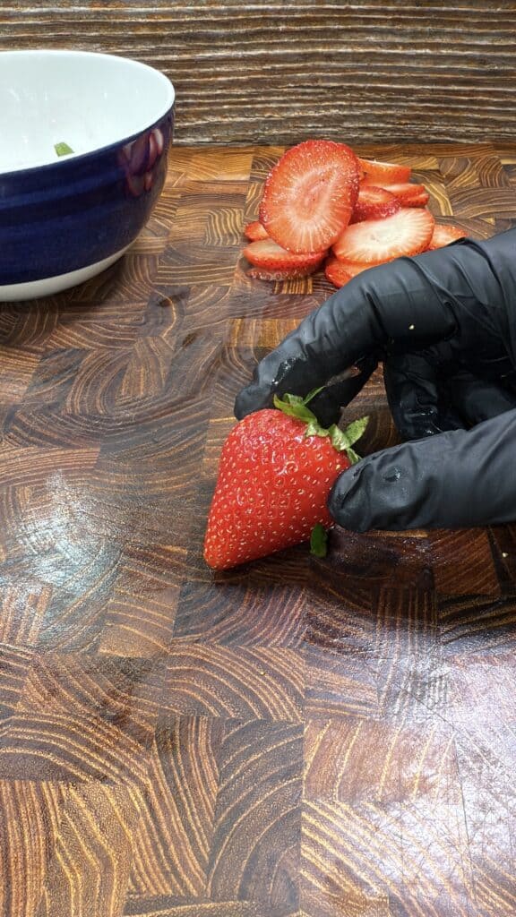 A hand wearing a black glove holds a whole strawberry on a wooden cutting board, with sliced strawberries and a blue bowl in the background.