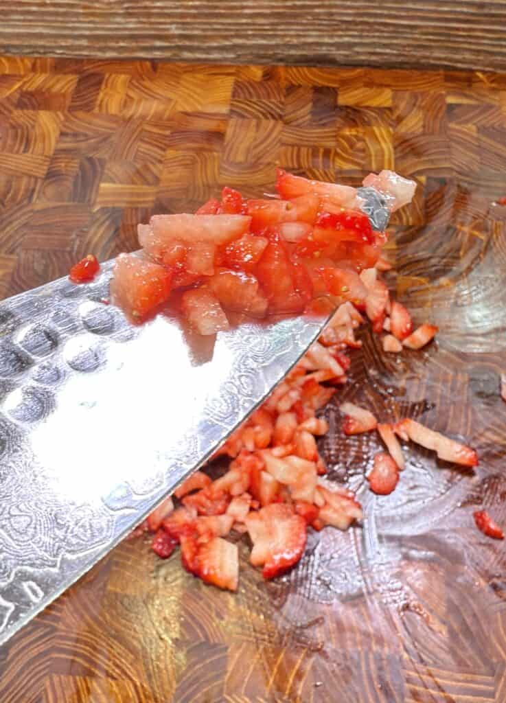 A close-up of finely chopped strawberries on a patterned knife blade, with more diced strawberries scattered on a wooden cutting board in the background.