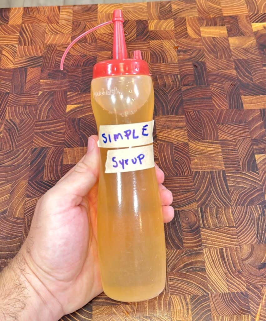 A hand holding a clear squeeze bottle with a red cap, containing a light yellow liquid labeled SIMPLE SYRUP in handwritten blue letters on masking tape. The background shows a wooden cutting board with a checkered pattern.