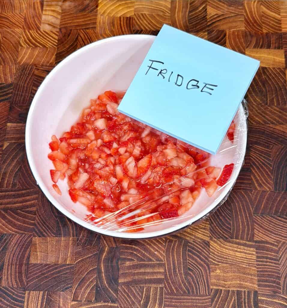 A white bowl filled with chopped strawberries covered in plastic wrap sits on a wooden surface. A blue sticky note labeled FRIDGE is placed on top.