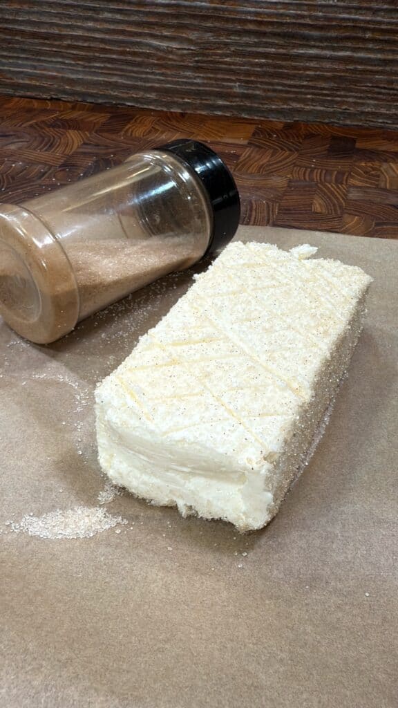 A rectangular block of white cheese with a crosshatch pattern is sprinkled with a seasoning, shown spilling from a clear plastic shaker, on a piece of parchment paper atop a wooden surface.