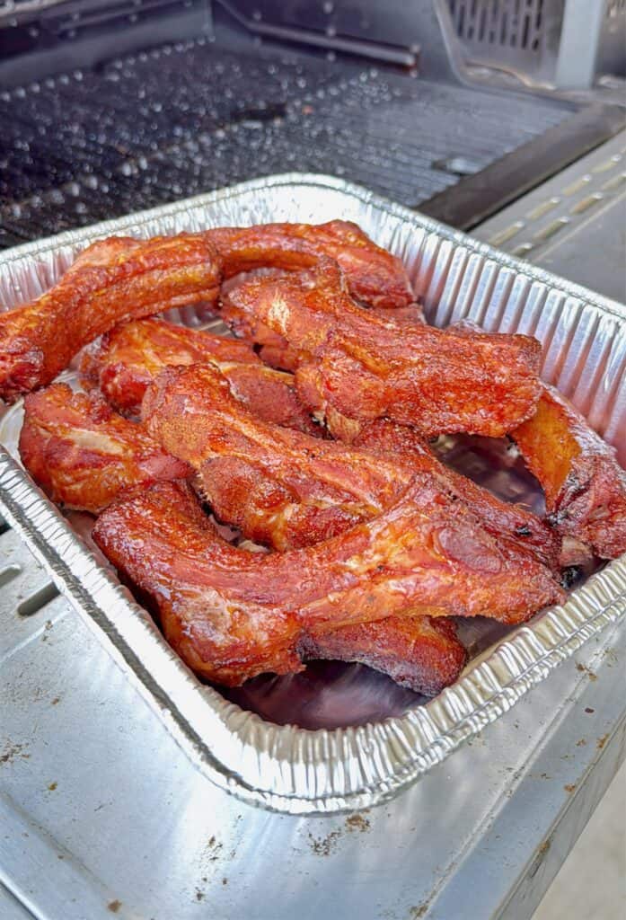 A foil tray filled with several pieces of grilled, seasoned pork ribs sits on the side of an outdoor barbecue grill. The ribs appear crispy and well-cooked with a reddish-brown color.