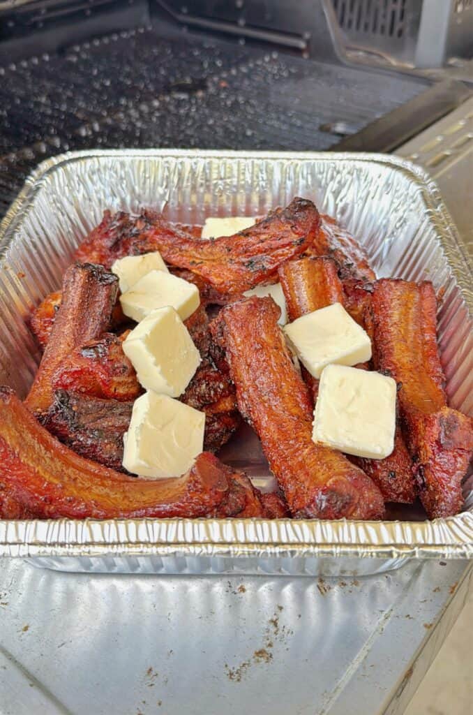 A foil tray filled with grilled pork belly burnt ends, topped with several cubes of butter, sits on a grill. The meat is caramelized and richly colored.