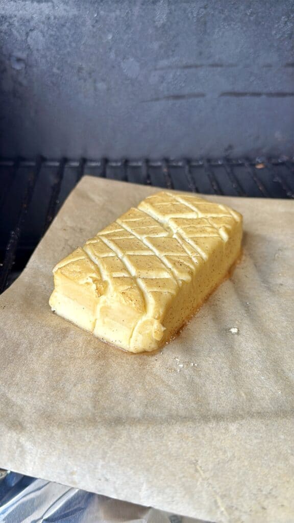 A rectangular block of soft cheese with a crisscross pattern on top rests on parchment paper, inside a grill or oven, ready to be cooked.