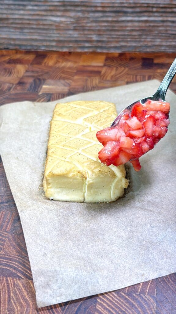 A rectangular cheesecake rests on parchment paper, while a spoonful of chopped strawberries is held above it, ready to be added as a topping. The background features a wooden surface.