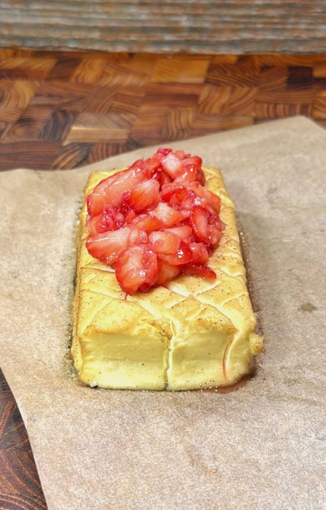 A loaf of baked tofu topped with chopped strawberries sits on parchment paper on a wooden surface. The tofu has a crisscross pattern on top and is garnished with juicy, fresh strawberries.