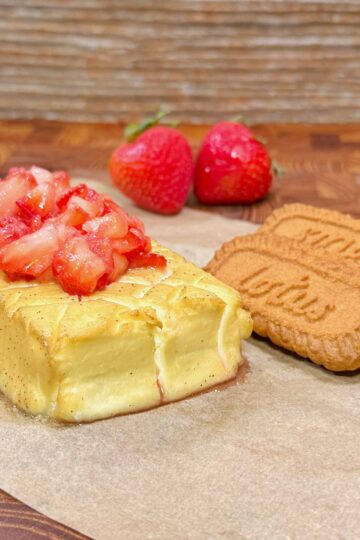 A rectangular cheesecake topped with chopped strawberries sits on parchment paper next to three Lotus Biscoff cookies and two whole strawberries, with a wooden surface background.