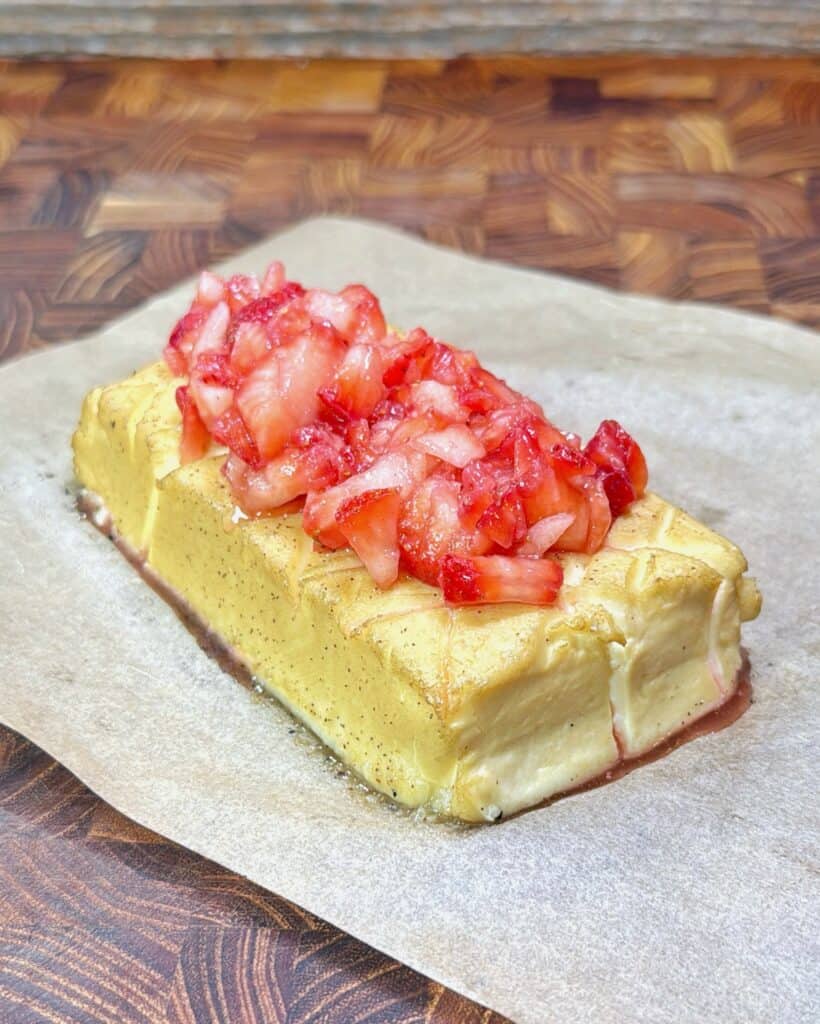 A rectangular block of cheesecake topped with chopped strawberries sits on a sheet of parchment paper atop a wooden surface. Recipe is of Strawberry Shortcake Smoked Cream Cheese