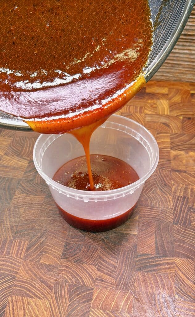 Thick reddish-brown sauce is being poured from a pan into a clear plastic container on a wooden countertop.