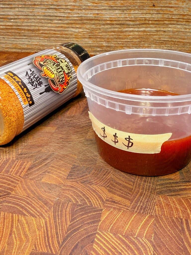 A plastic cup filled with reddish-brown sauce sits on a wooden surface next to a bottle of orange seasoning. The cup has a label with hand-drawn dollar signs taped to its side.