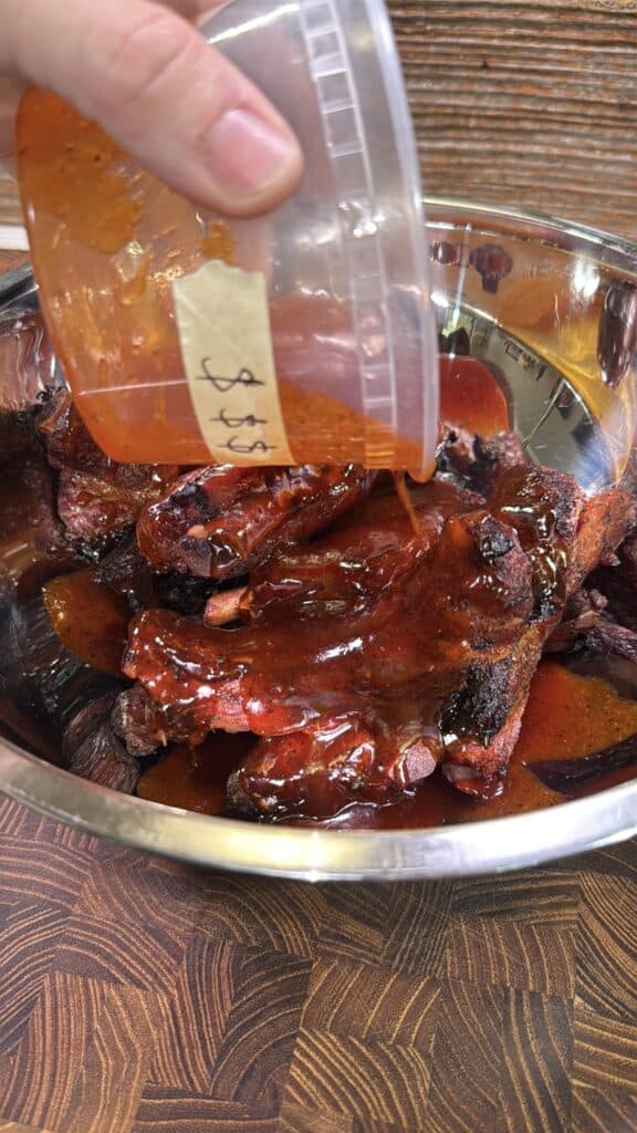 A hand pours a reddish barbecue sauce from a plastic container onto a bowl of cooked, charred ribs sitting on a wooden surface.