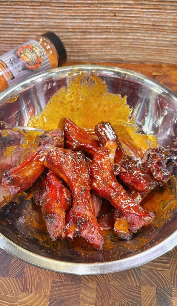 A metal bowl filled with glazed, grilled meat drumsticks coated in a reddish sauce, with a seasoning bottle lying on its side in the background on a wooden surface.