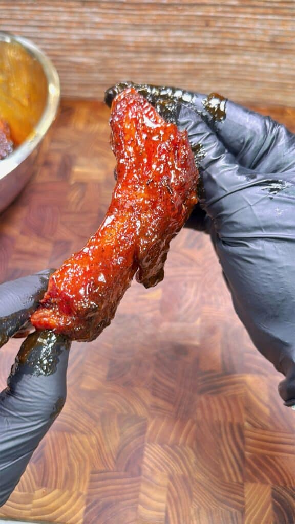 A person wearing black gloves holds a piece of saucy, red, glazed meat over a wooden surface, with a metal bowl partially visible in the background.