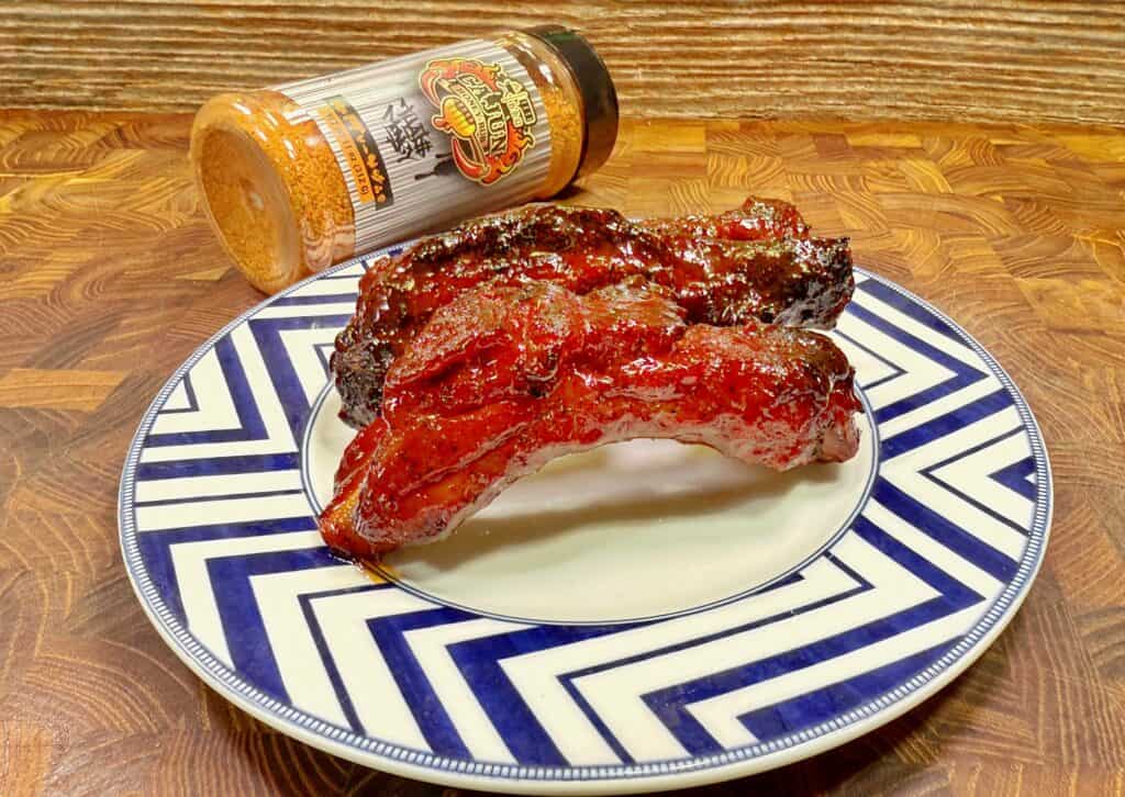 A plate with two glazed, barbecued ribs sits on a blue and white zigzag-patterned plate. Behind the plate is a bottle of barbecue seasoning tipped on its side on a wooden surface. Recipe is for Cajun Honey Party Ribs.