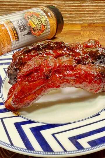 A plate with two glazed, barbecued ribs sits on a blue and white zigzag-patterned plate. Behind the plate is a bottle of barbecue seasoning tipped on its side on a wooden surface.