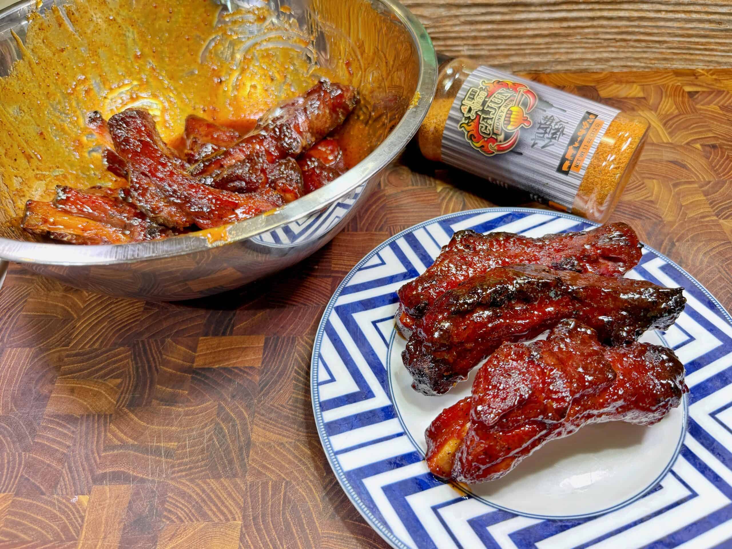 A plate with glazed, grilled pork ribs sits on a geometric-patterned dish beside a bowl containing more ribs and a spice bottle on a wooden surface. The ribs look caramelized and sticky.