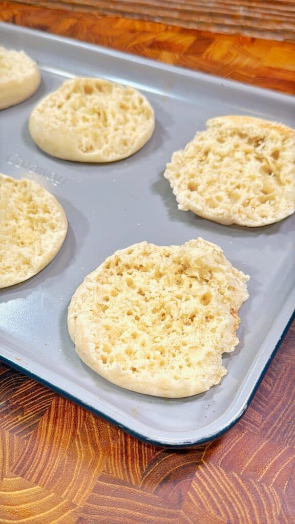 Four English muffin halves with a golden, airy texture sit on a gray baking sheet placed on a wooden surface. The muffins have a rough, porous appearance typical of traditional English muffins.