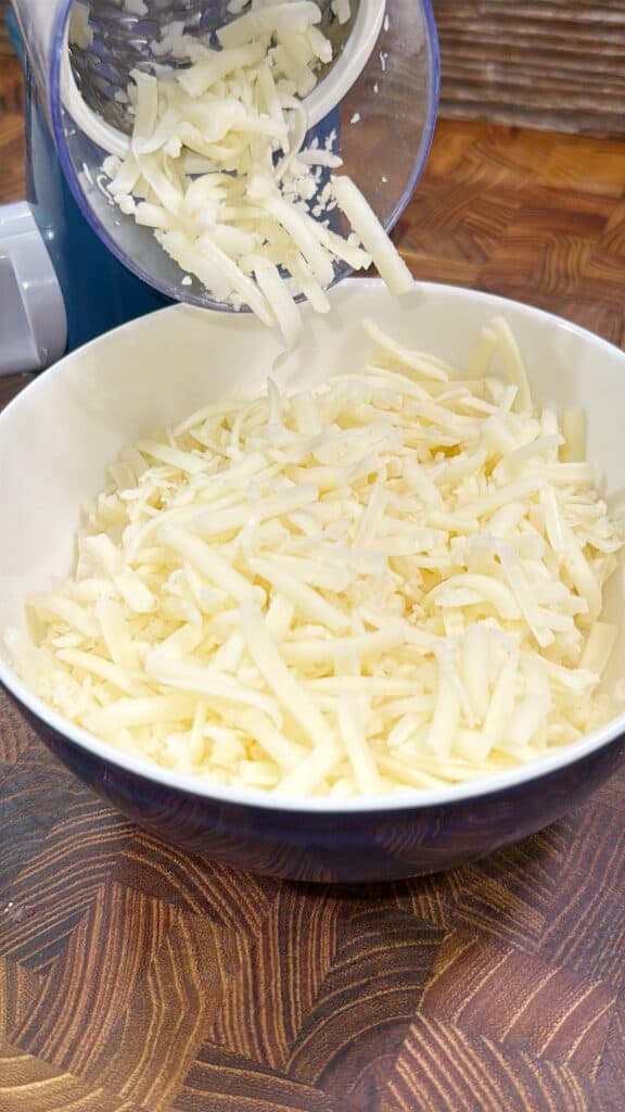A bowl filled with freshly grated white cheese sits on a wooden countertop, with more cheese being grated into the bowl from a kitchen grater above.