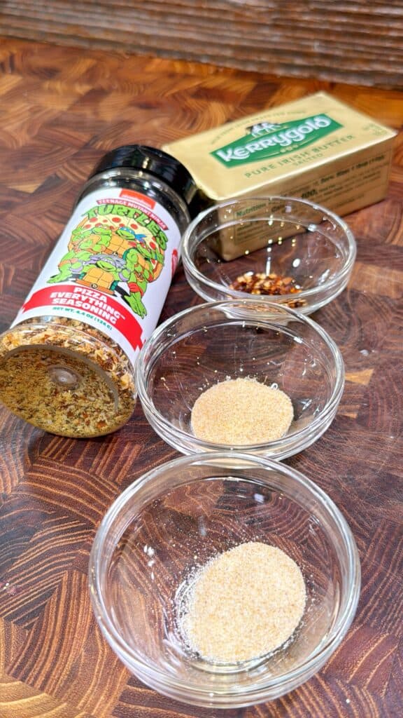A bottle of seasoning, a stick of Kerrygold butter, red pepper flakes, onion powder, and garlic powder are arranged in small glass bowls on a wooden cutting board.