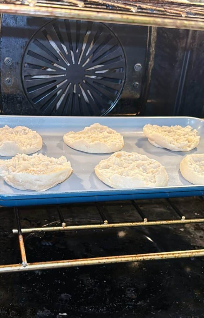 Six halved English muffins are toasting on a blue baking tray inside an oven. The muffins are spaced apart, with their cut sides facing up. The oven’s fan and interior are visible in the background.
