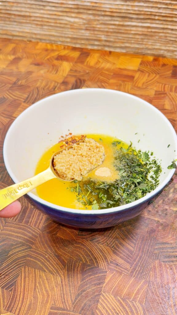 A white bowl with melted butter, dried herbs, and a teaspoon of minced garlic sits on a wooden surface. The ingredients are being combined for a recipe.