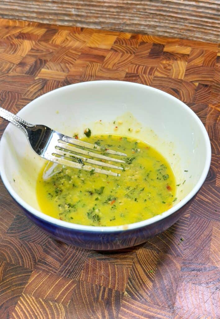 A white bowl with a blue exterior contains a yellow-green herb vinaigrette. A fork rests inside the bowl, which sits on a wooden patterned cutting board.